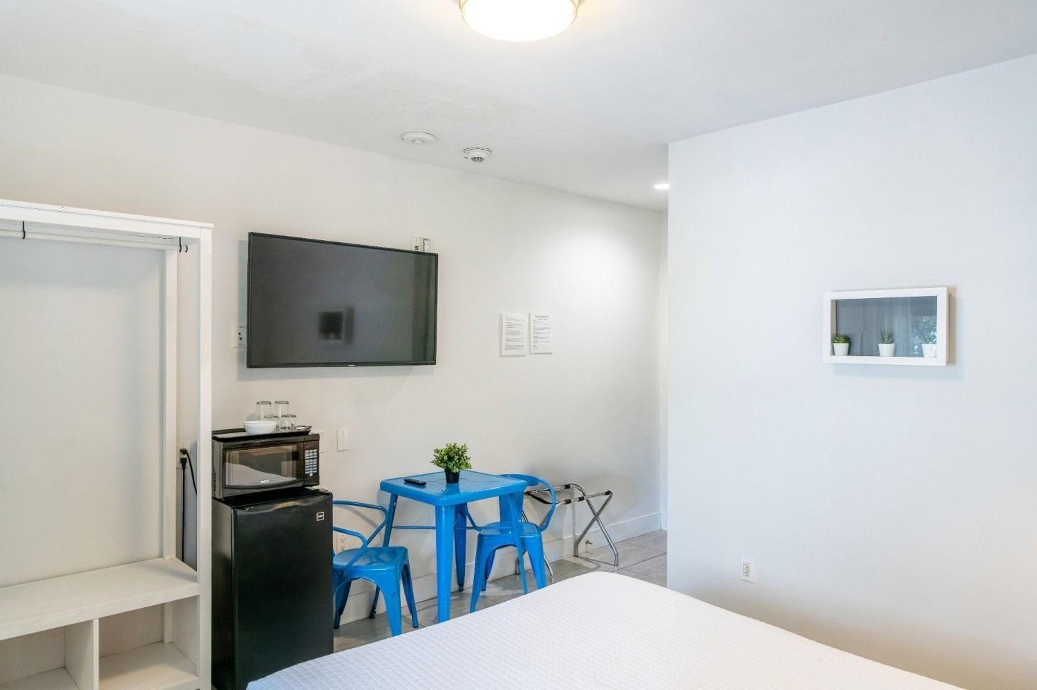 Small hotel room with a mounted TV, a mini-fridge, and blue table and chairs.