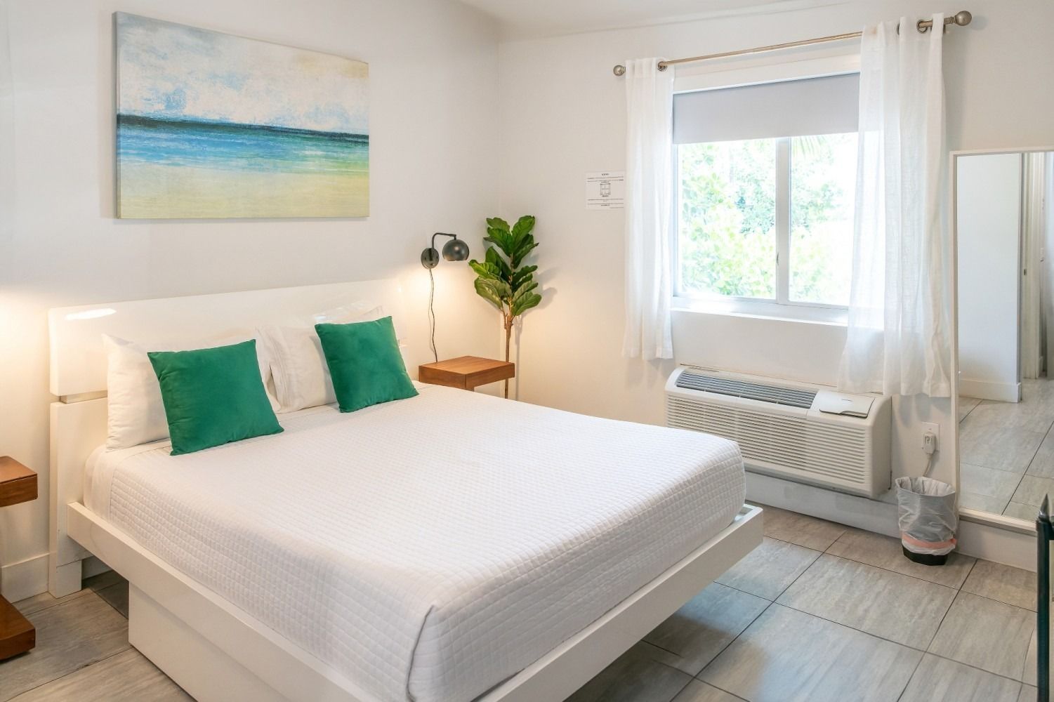White-walled bedroom with white bed, green pillows, beach art, window, and small plant.