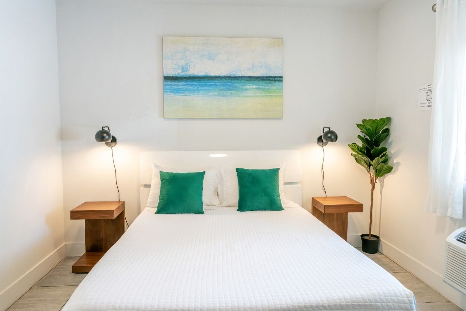 Bedroom with white walls, bed, wooden bedside tables, artwork, and a potted plant.