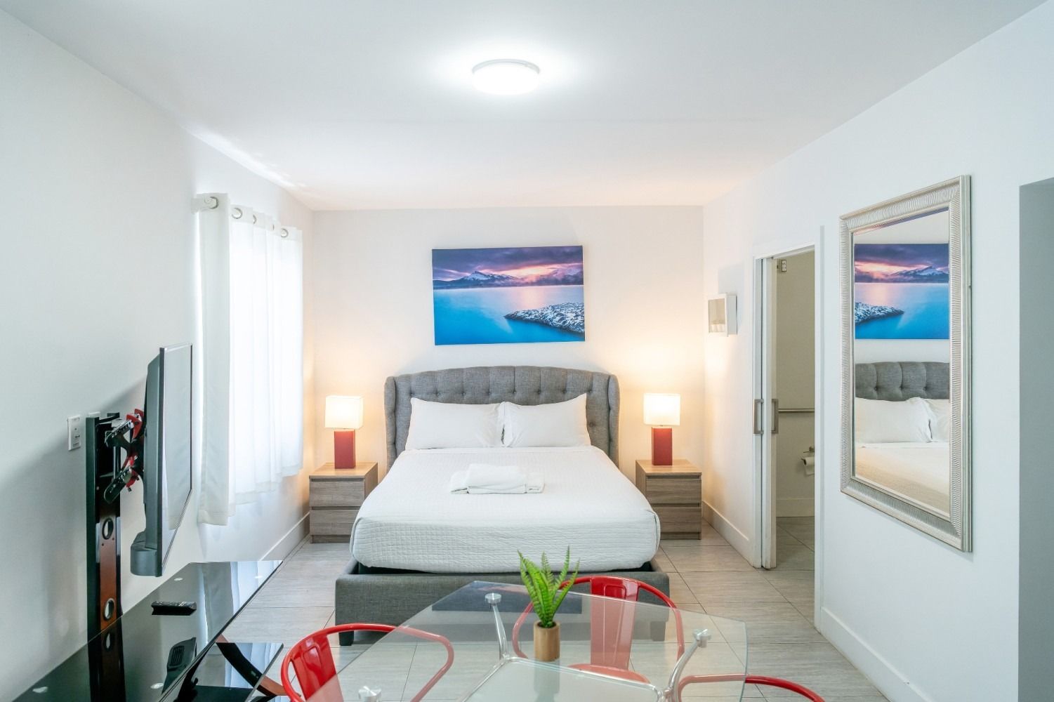 Bedroom with a bed, nightstands, TV, table, and a bathroom. Neutral tones and red accents.