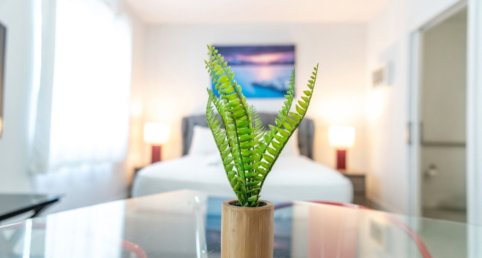 Green plant in a wooden pot on a glass table in front of a bed.