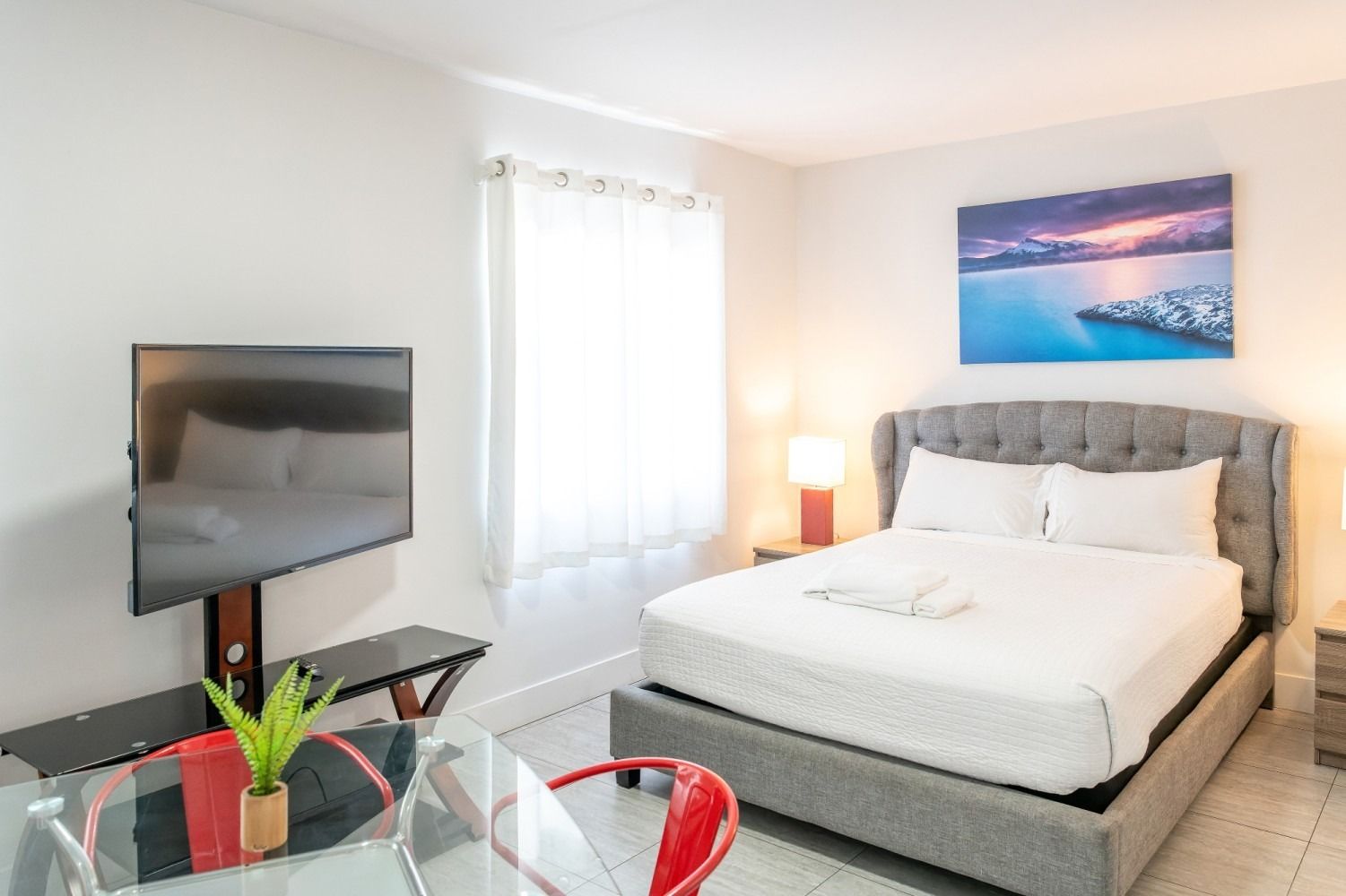 Bedroom with a gray bed, large TV, small table, red chair, and a window.