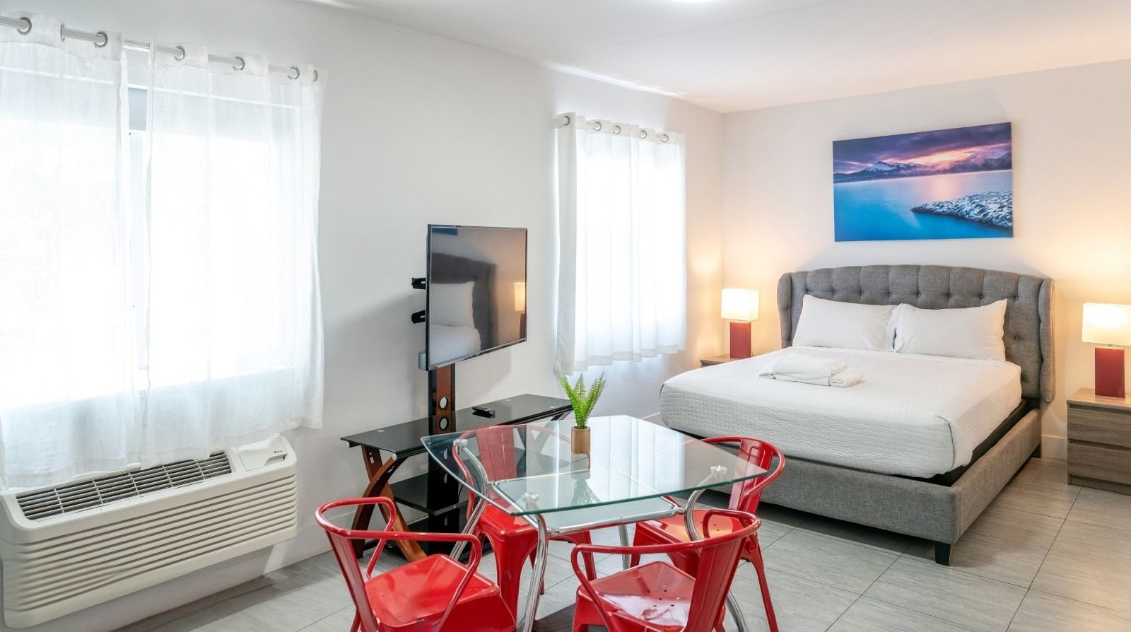 Studio apartment with a queen bed, dining table, and TV. Bright white walls and red accents.