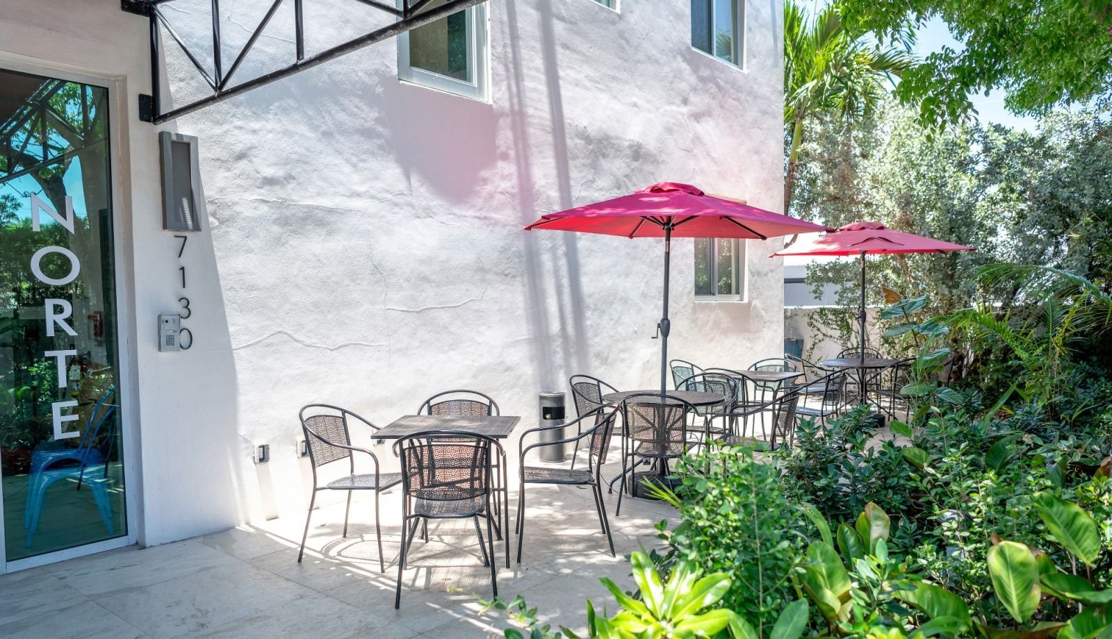 Outdoor cafe with tables, chairs, and pink umbrellas next to a white building.