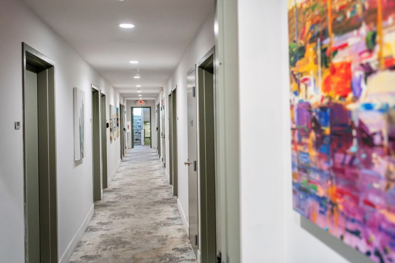 Long hallway with multiple doors, neutral colors, and artwork. Carpeted floor, overhead lighting.