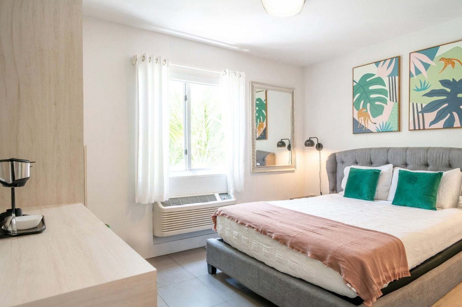 Bedroom with a gray bed, white walls, two green pillows, pink throw, and botanical art.