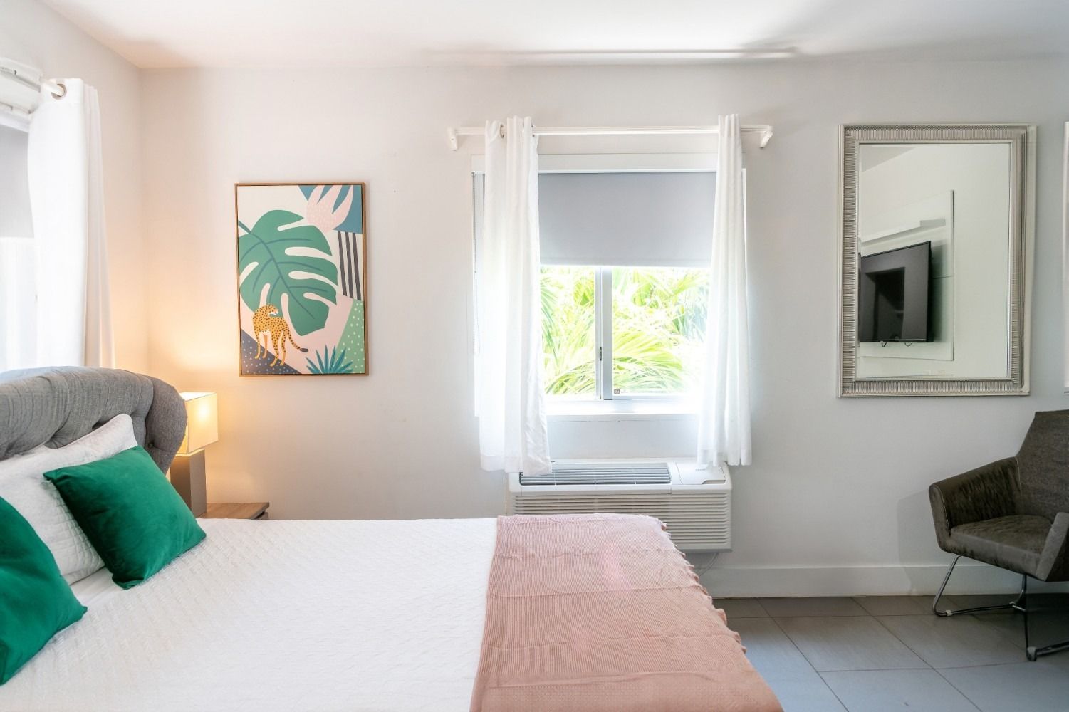 Bedroom with a bed, artwork, window, and armchair. Bright and airy decor with green and pink accents.