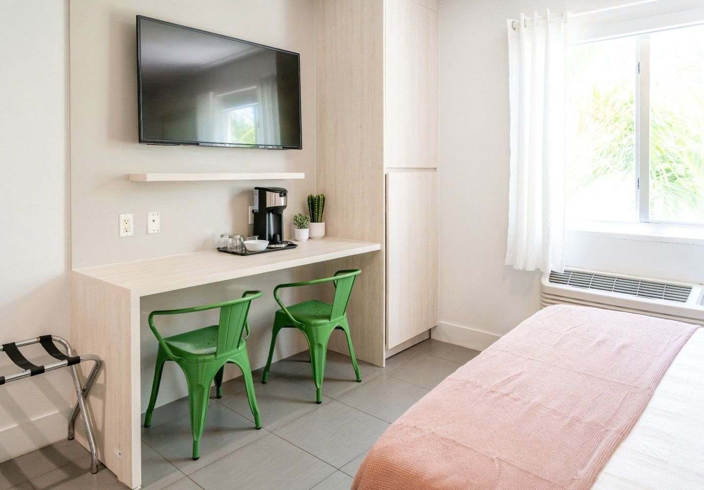 Hotel room with a bed, desk, and TV. Green chairs, white walls, and a window.