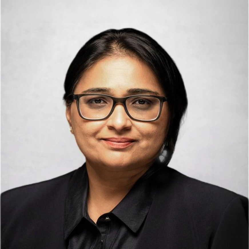 Professional headshot of a person wearing black glasses and a black blazer, posed against a light gray background.