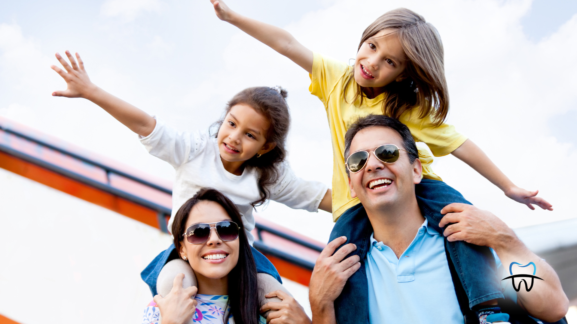 Family with two children on parents' shoulders, arms outstretched, smiling. Outdoors, sunny day.