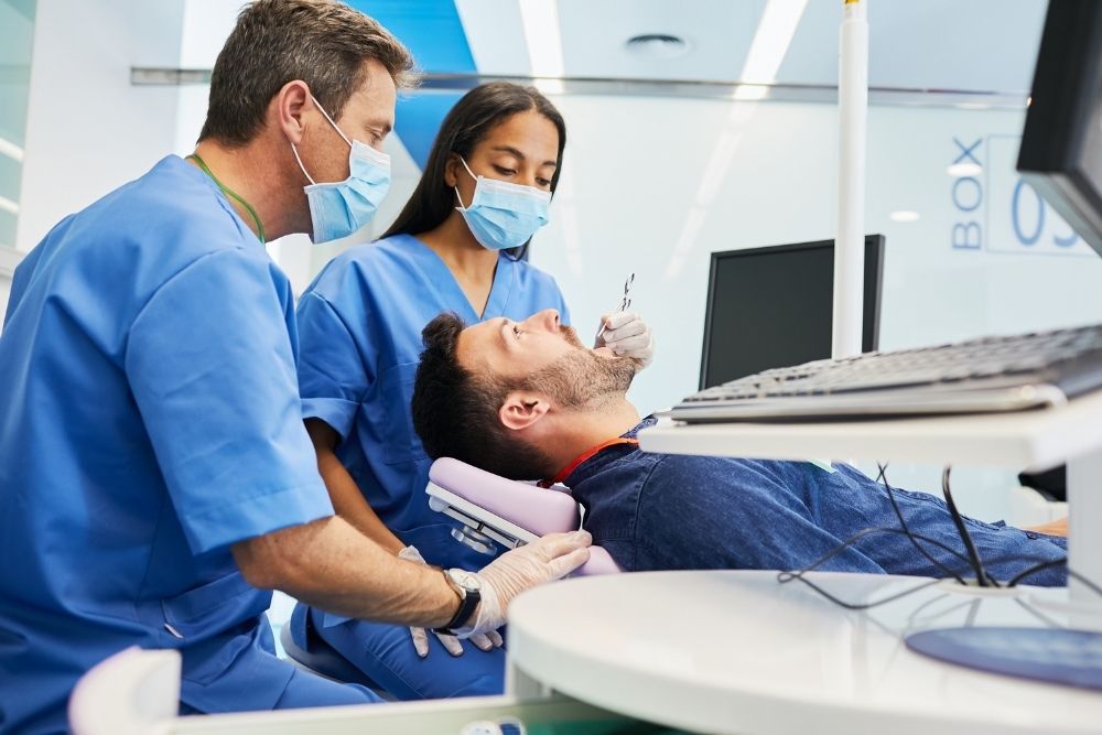 Dentist and assistant examining a patient's teeth in a dental clinic; all are wearing face masks.