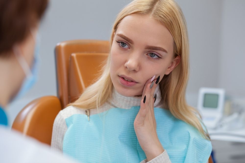Woman in dental chair, holding cheek, speaking to dentist wearing a mask.