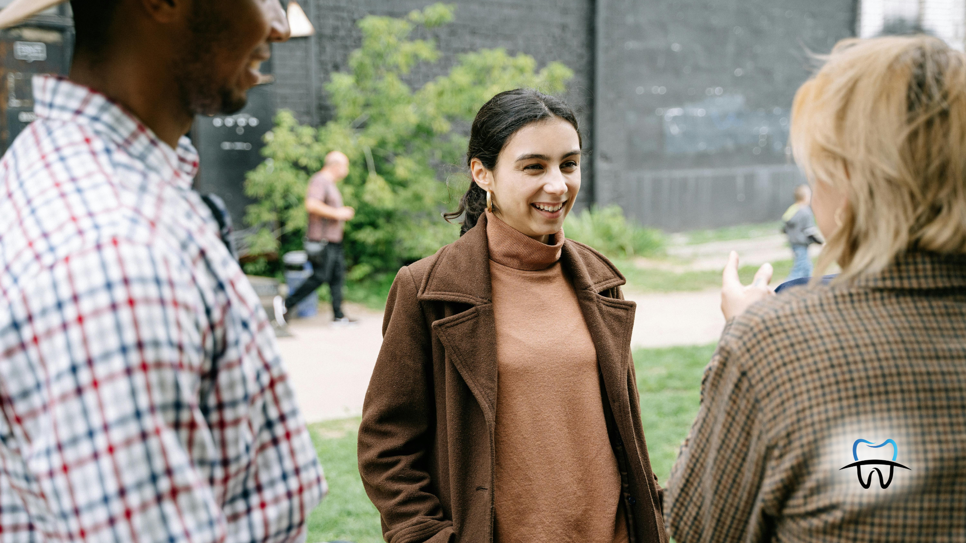 Three people conversing outdoors; woman in brown coat smiles, two others face her.