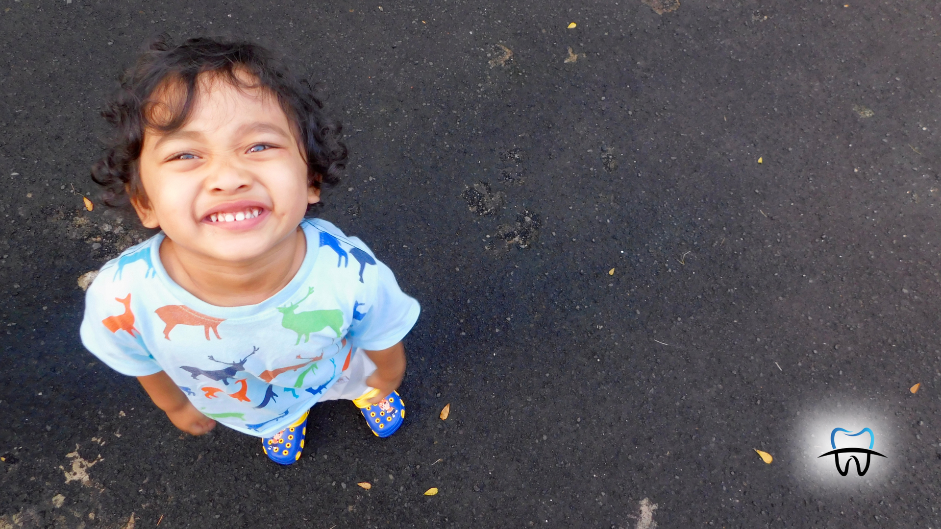 Smiling child looking upwards, standing on a dark, textured surface, wearing a blue shirt with animal prints.