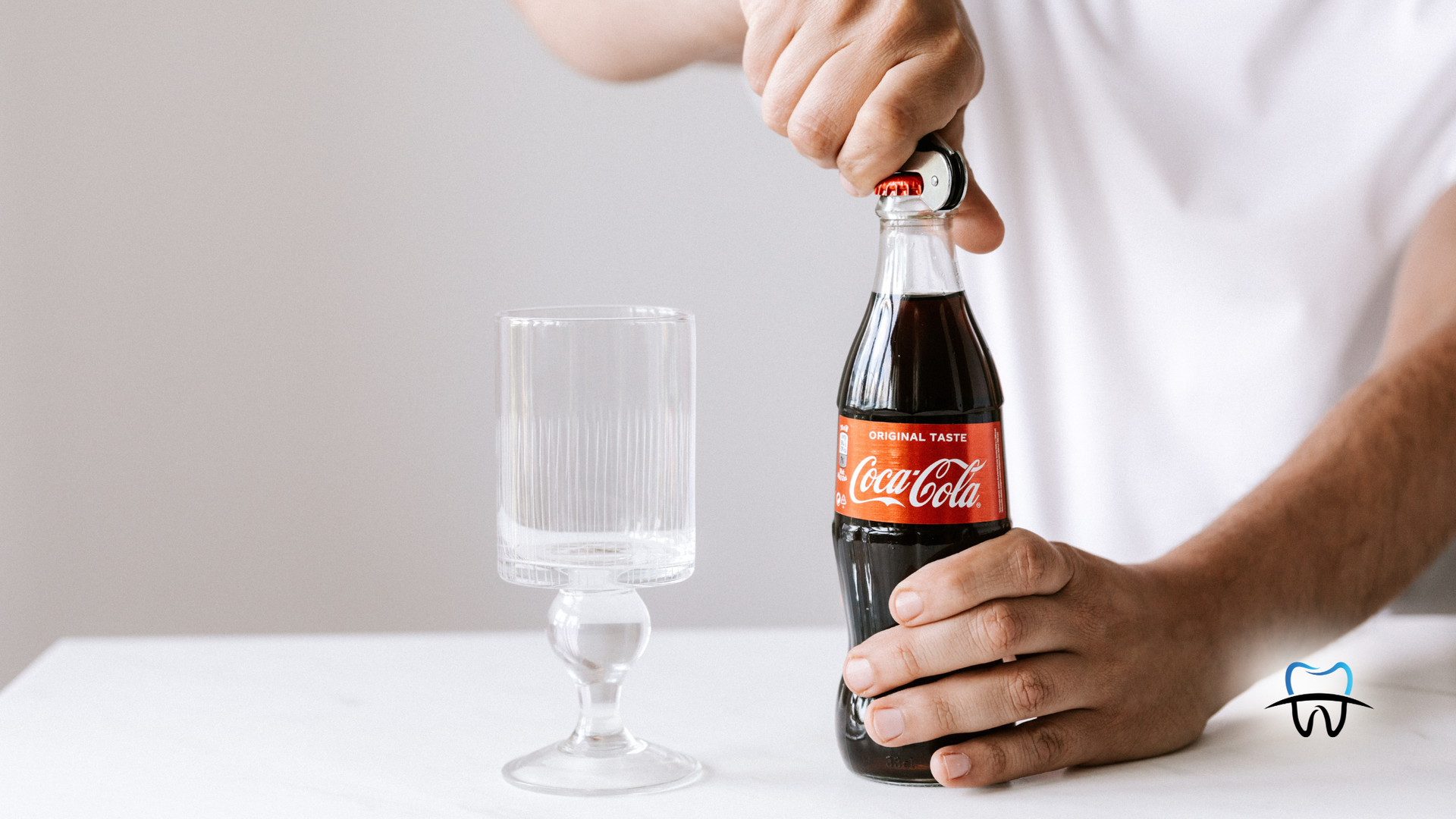 Person opening a Coca-Cola bottle with a glass next to it on a white table.