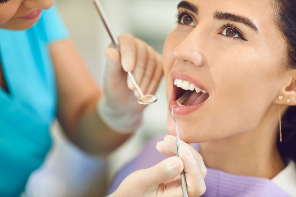 Woman at dentist with mouth open; tools held by gloved hands.