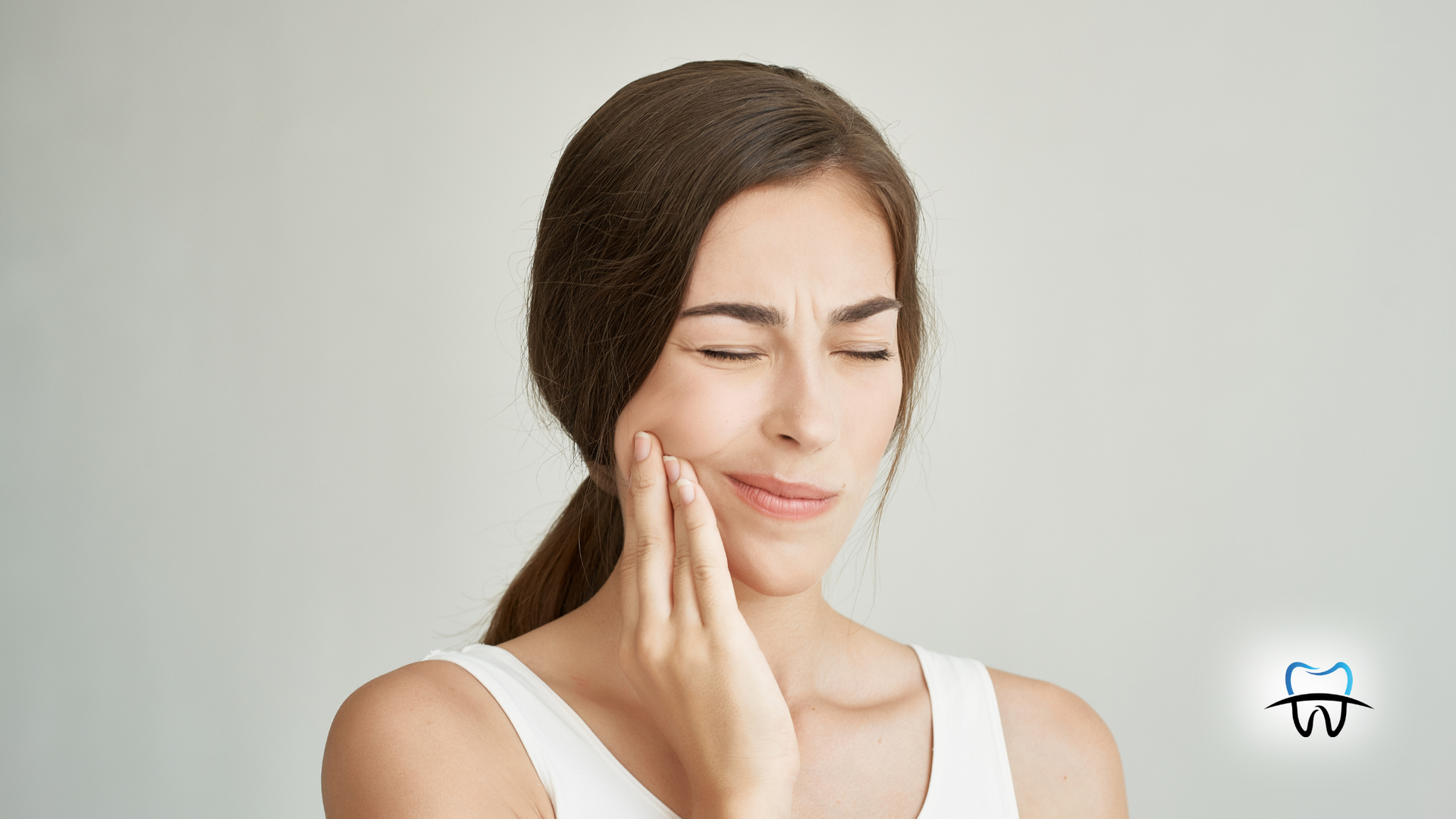 Woman with a hand on her cheek, eyes closed, appearing to be in pain against a light background.