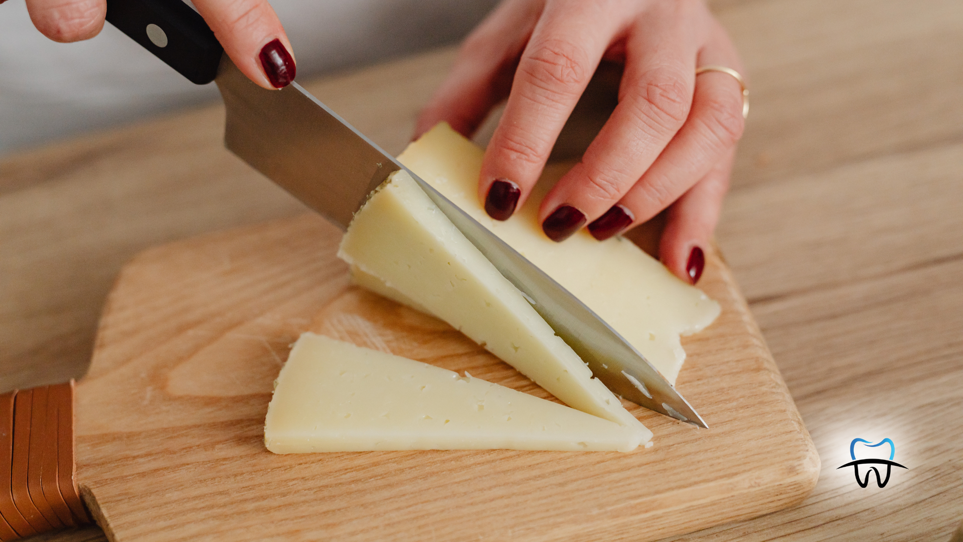Hands with maroon nails slice cheese on a wooden cutting board with a knife.