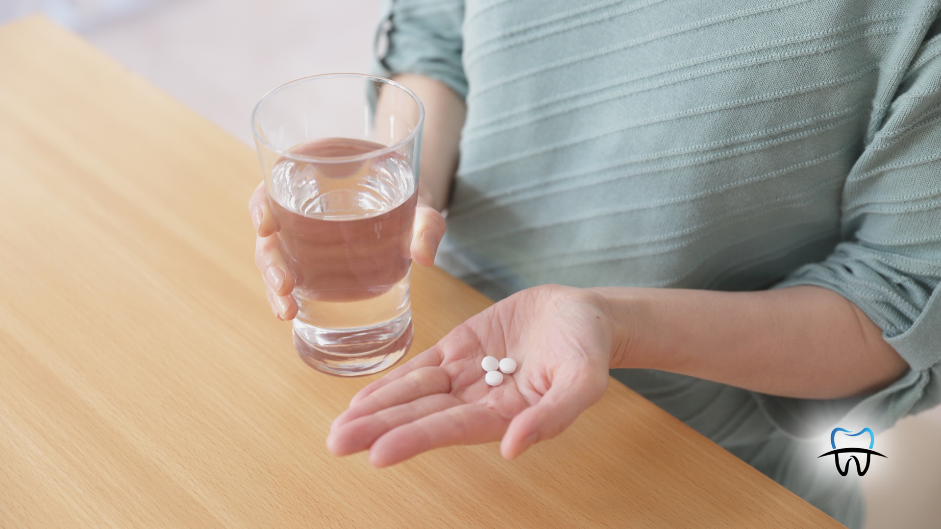 Person holding pills in one hand and a glass of water in the other, seated at a wooden table.
