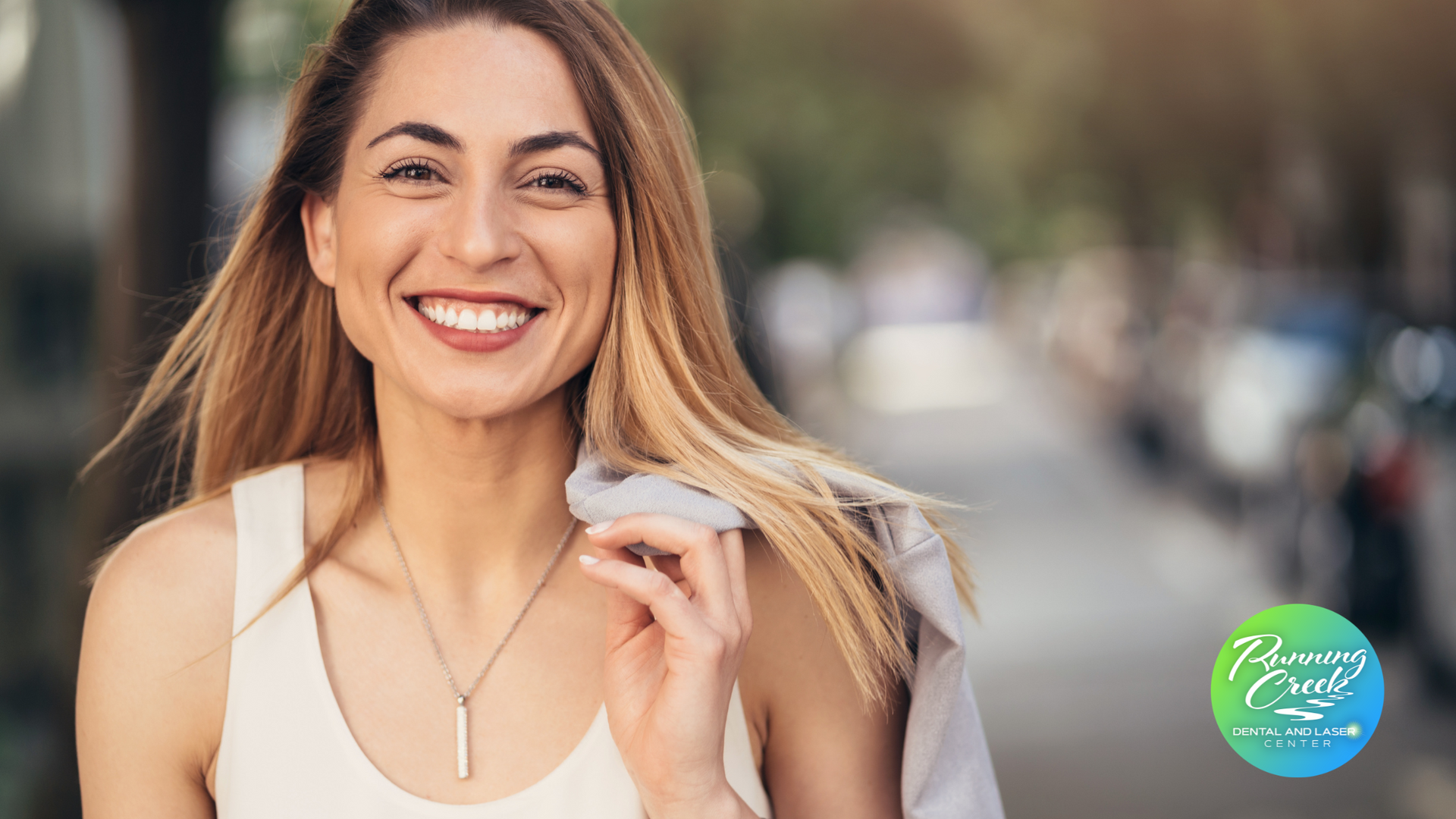 A woman wearing a white tank top and a necklace is smiling.