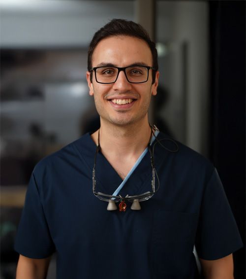 Smiling person in black scrubs in a dental office, loupes around neck.