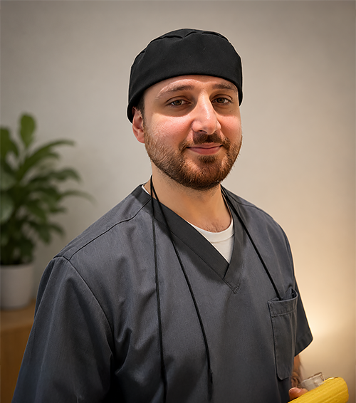 Smiling person in black scrubs in a dental office, loupes around neck.