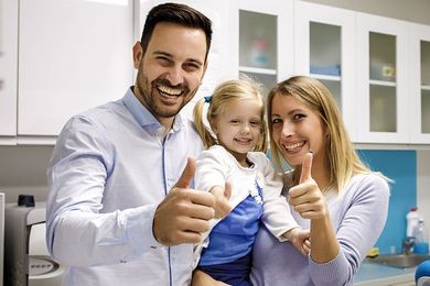 Family smiling and giving thumbs-up in a well-lit room with white cabinets; the woman holds a child.