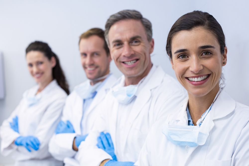 Four healthcare workers in white coats and blue gloves smiling, arms crossed.