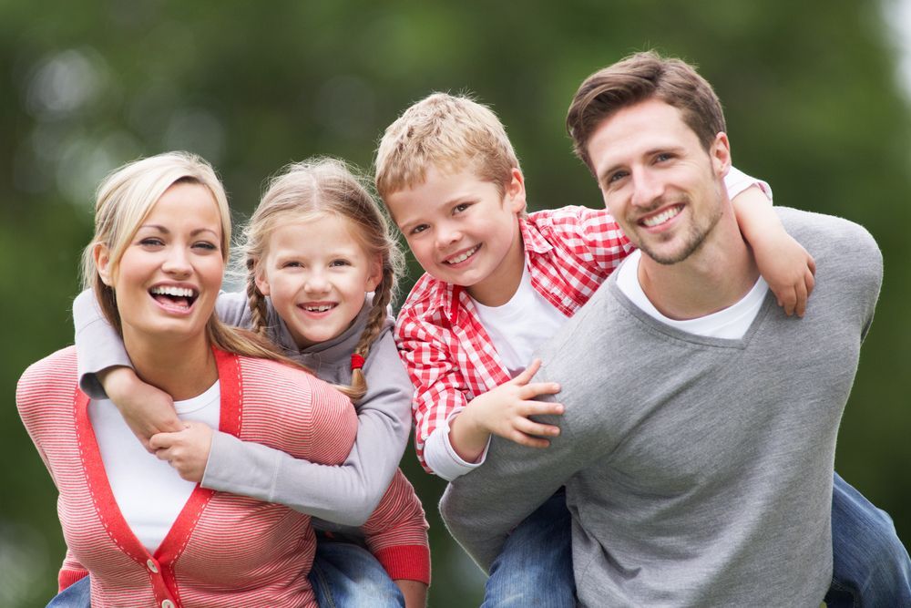 Family of four smiling, piggybacking each other outdoors.
