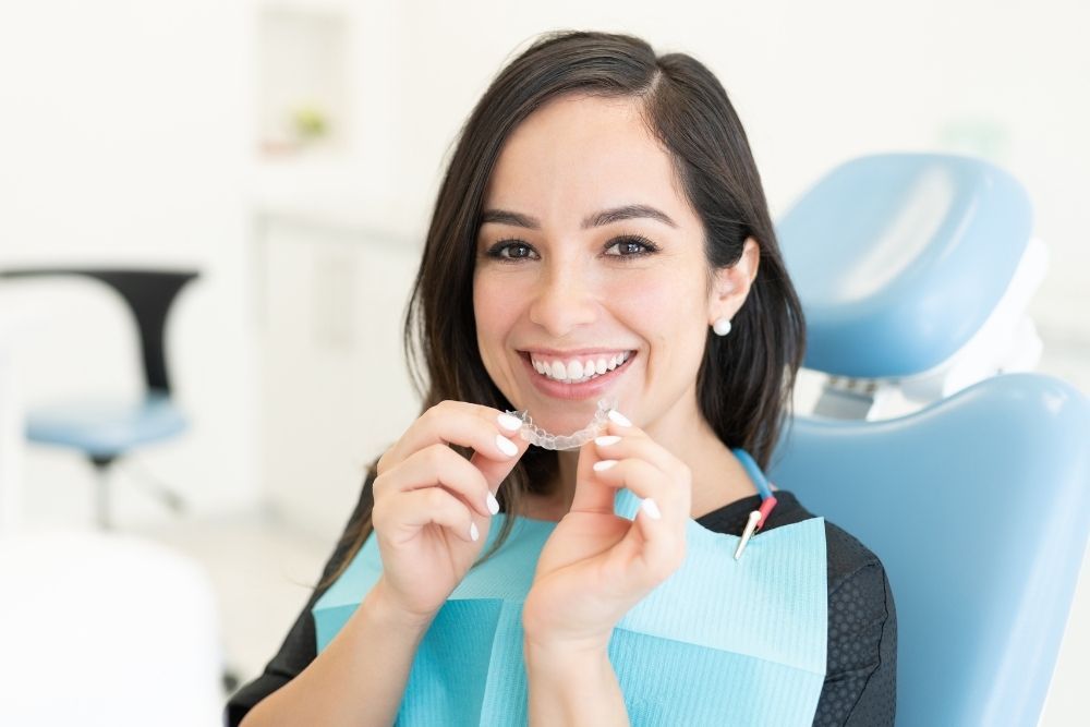 Woman in dental chair, holding clear aligner, smiling.