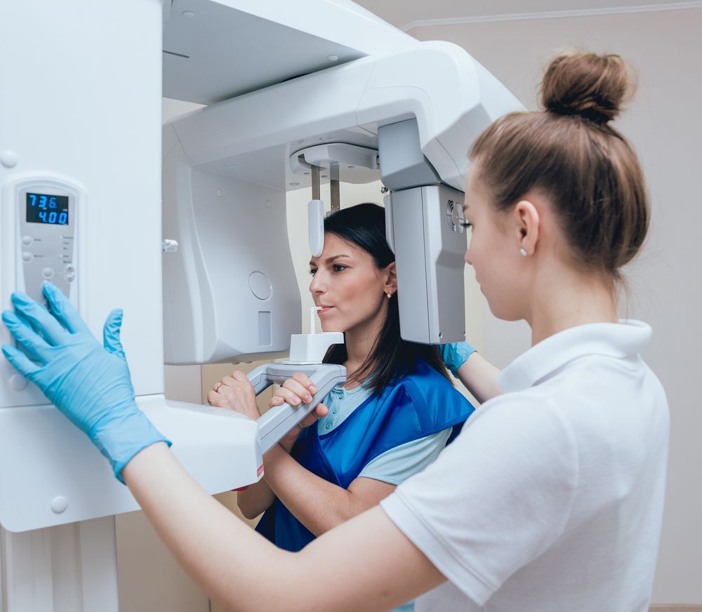 Woman undergoing dental X-ray. Dental assistant in blue gloves adjusts machine.
