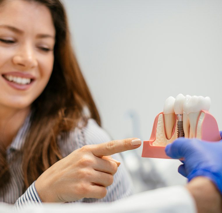 Woman smiling, pointing to dental implant model held by a person in blue gloves, dental office setting.