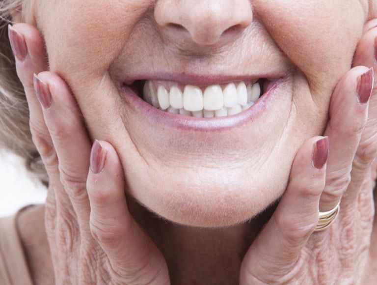 Woman's smiling mouth, hands on cheeks, showing white teeth, wrinkles visible, and manicured nails.