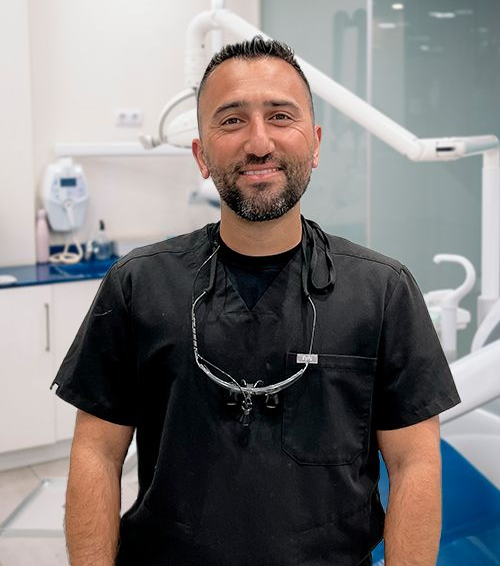 Dentist in black scrubs smiles in a dental clinic, glasses around his neck, equipment in background.