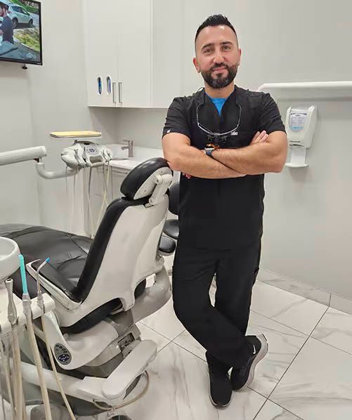 Dentist in scrubs, arms crossed, leaning against dental chair in a clean office.
