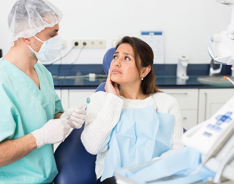 Dentist examining a patient with a hand on her cheek, expressing discomfort, in a dental office.