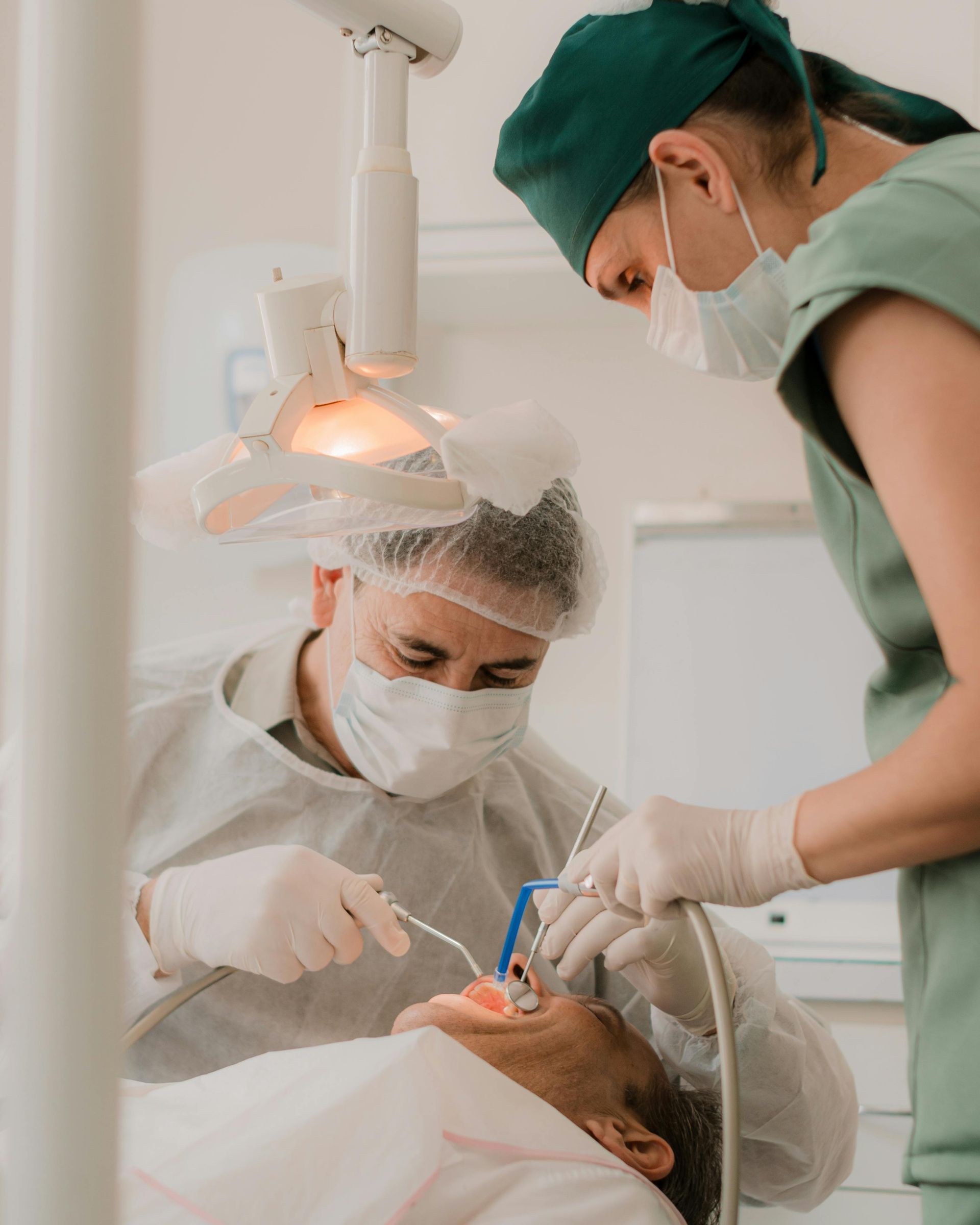 Dentist and assistant working on a patient in a dental office. Both wear masks, gowns, and gloves.