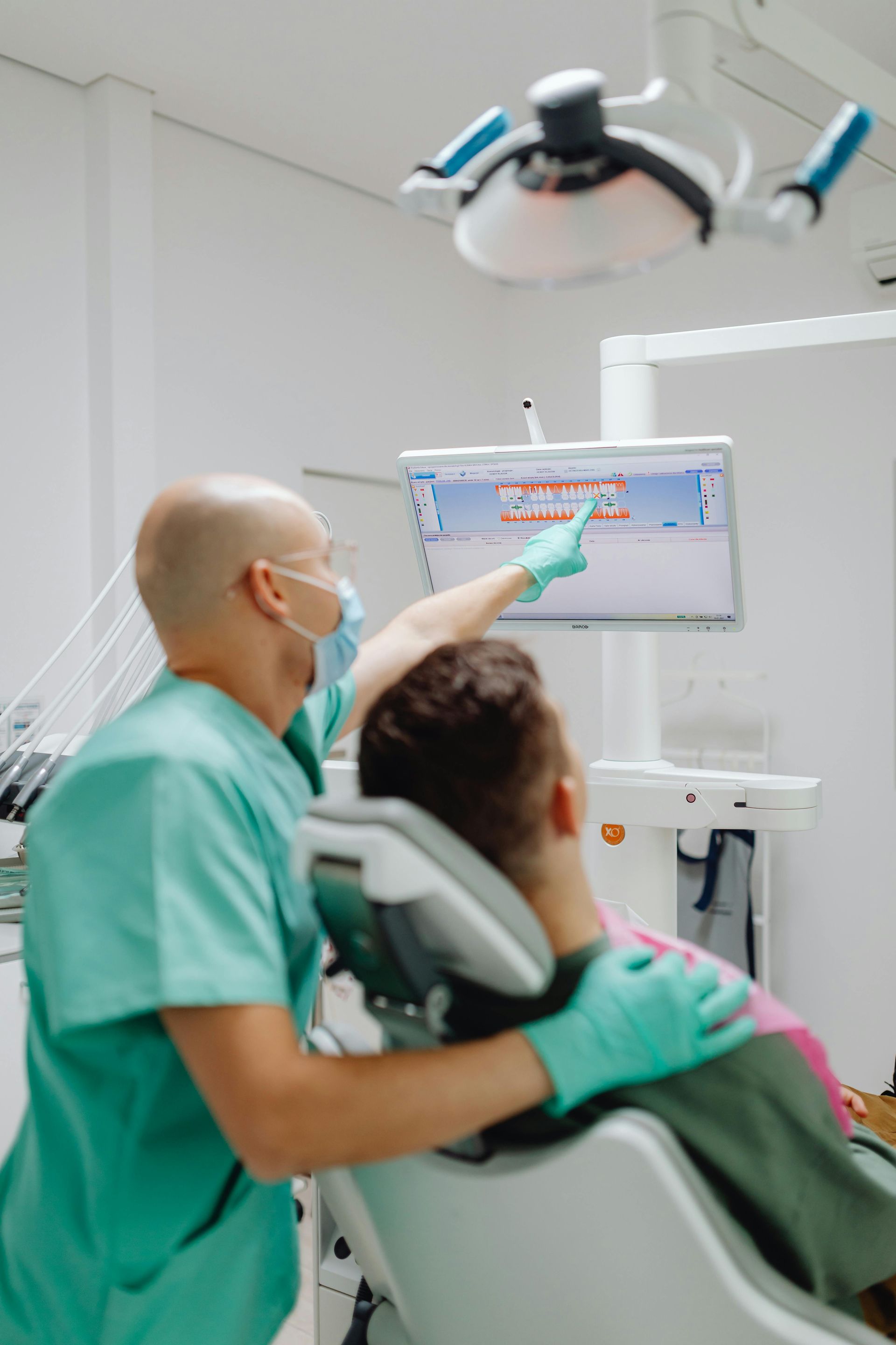 Dentist in green scrubs pointing at a screen with dental images, explaining to a patient in a dental chair.