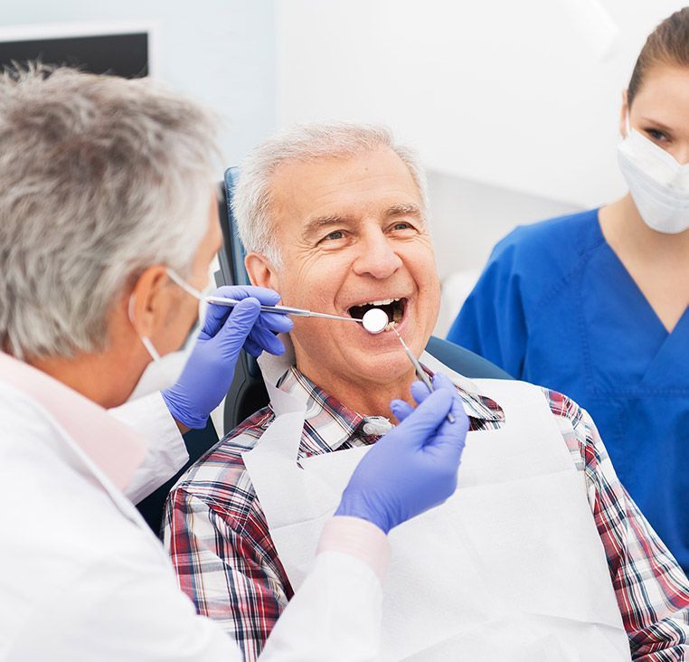 Dentist examining a patient's mouth with tools in an office, smiling. A dental assistant is nearby.