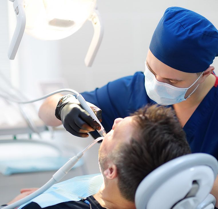 Dentist in blue scrubs, mask, and gloves, examining a patient's mouth under bright light.