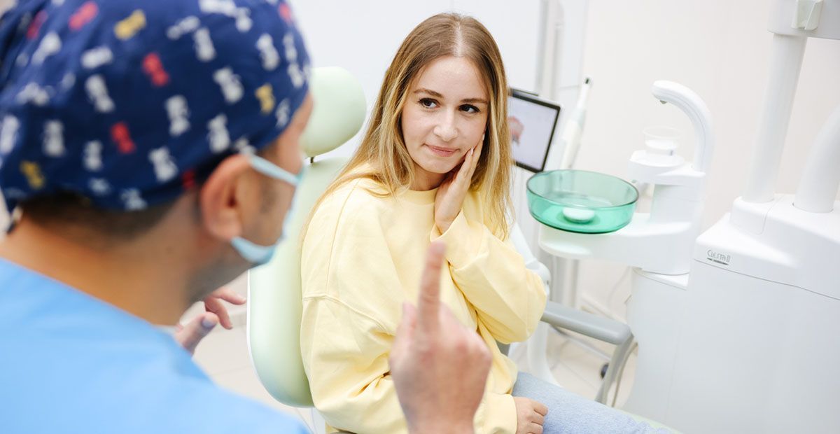 Woman in a dentist chair, holding her jaw and talking to the dentist.