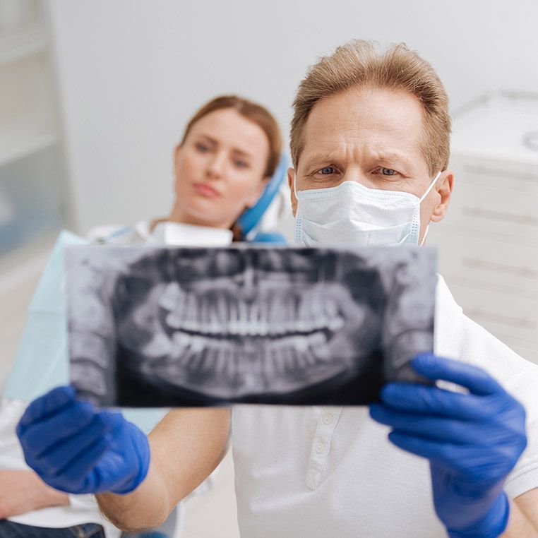 Dentist in mask and gloves examines dental x-ray; patient sits in chair behind him, looking concerned.