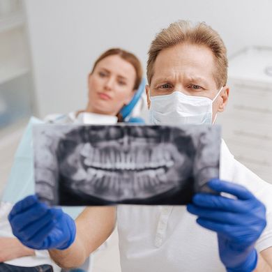 Dentist in mask and gloves examines dental X-ray, patient in background.