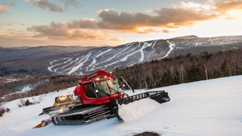 Snow groomers maintaining ski slopes at sunset, with a red machine pushing snow on a winter hillside.
