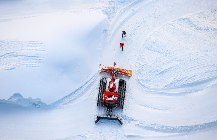 A snow grooming machine’s plow attachment, with its yellow blades and tracked wheels, resting on wooden planks.