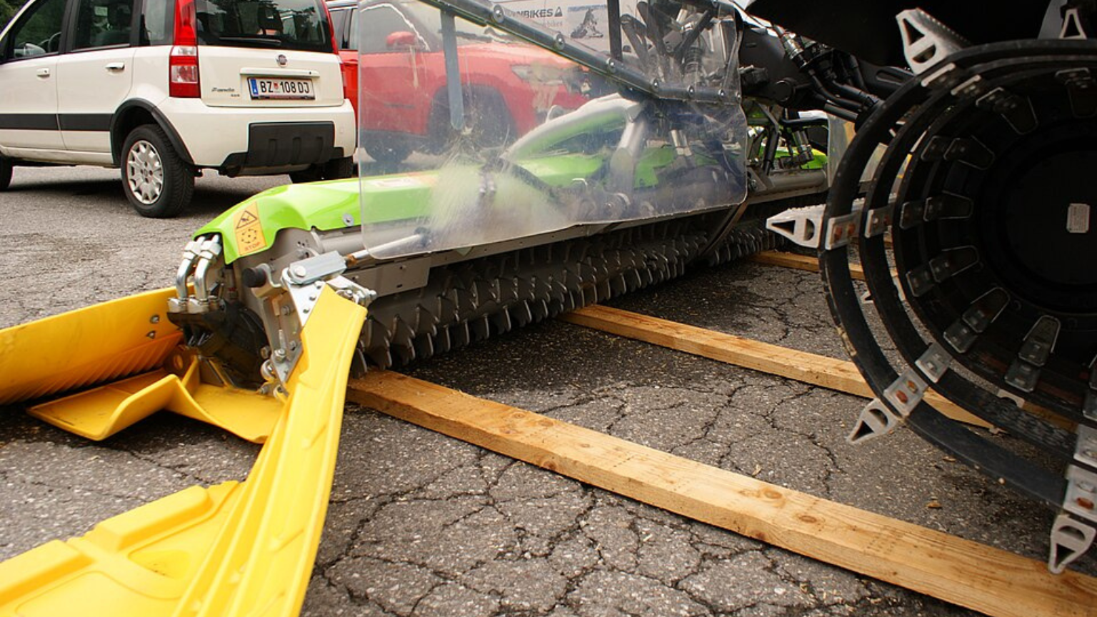A large snow groomer with wide tracks and snow plowing blades, parked on a snowy mountain slope, surrounded by trees.