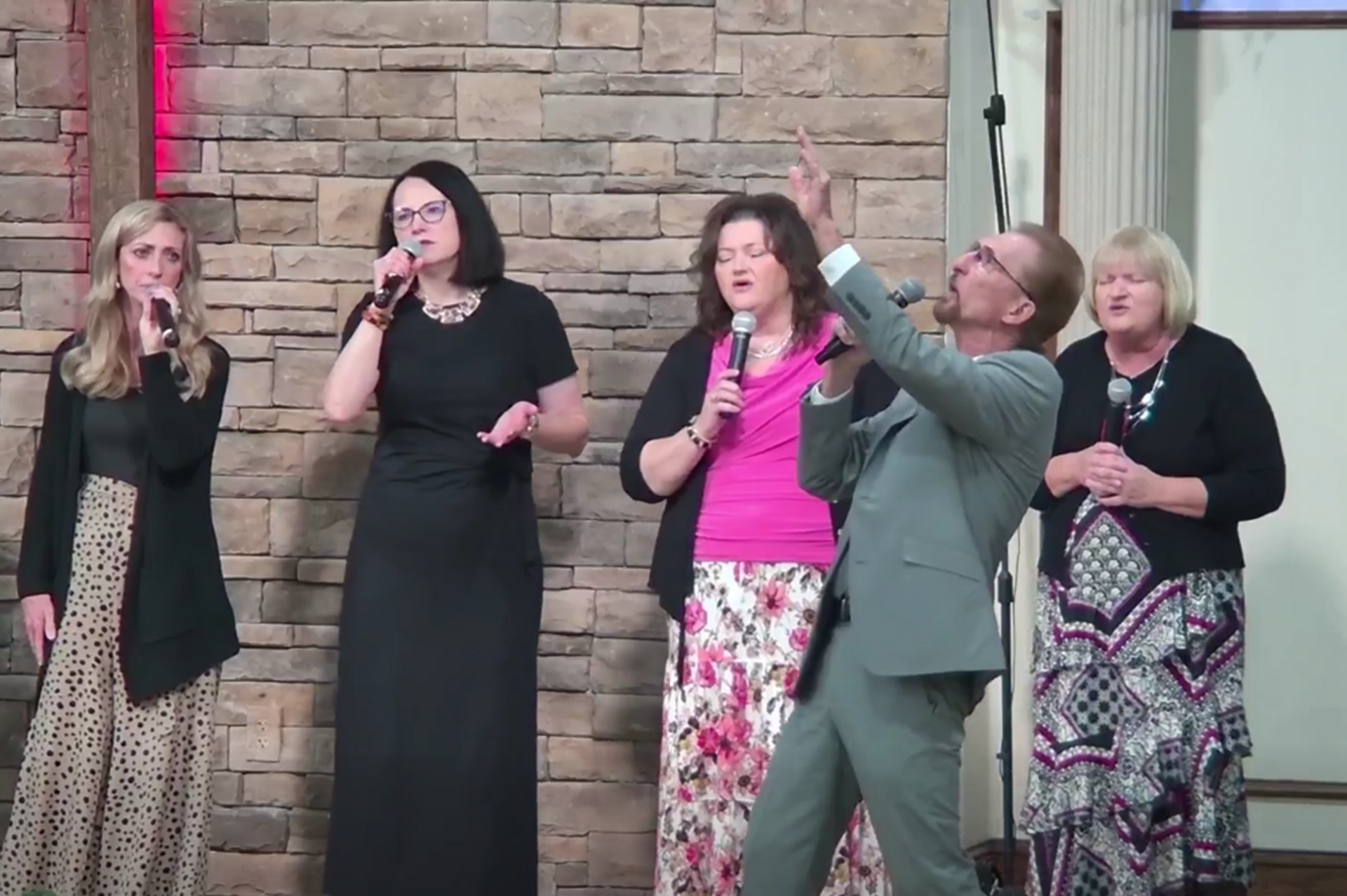 A group of people singing on a stage in front of a stone wall. One person points up.
