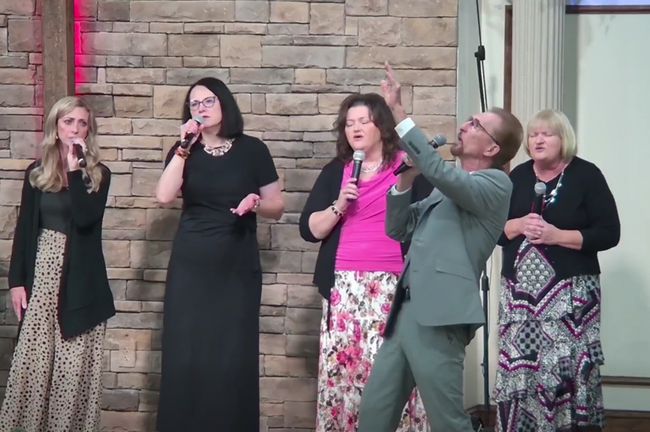 A group of people singing on a stage in front of a stone wall. One person points up.