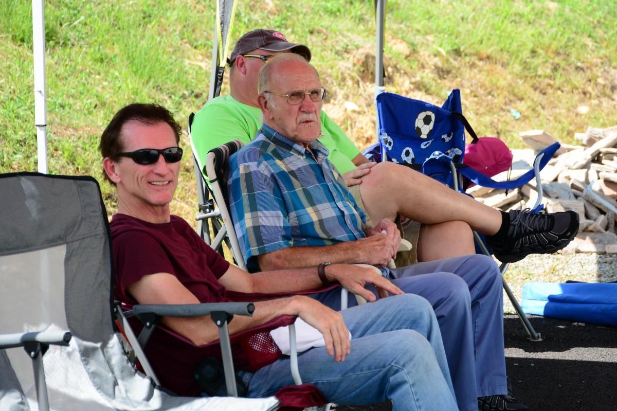 Three men in camp chairs under a canopy; one wearing sunglasses, another in a plaid shirt.