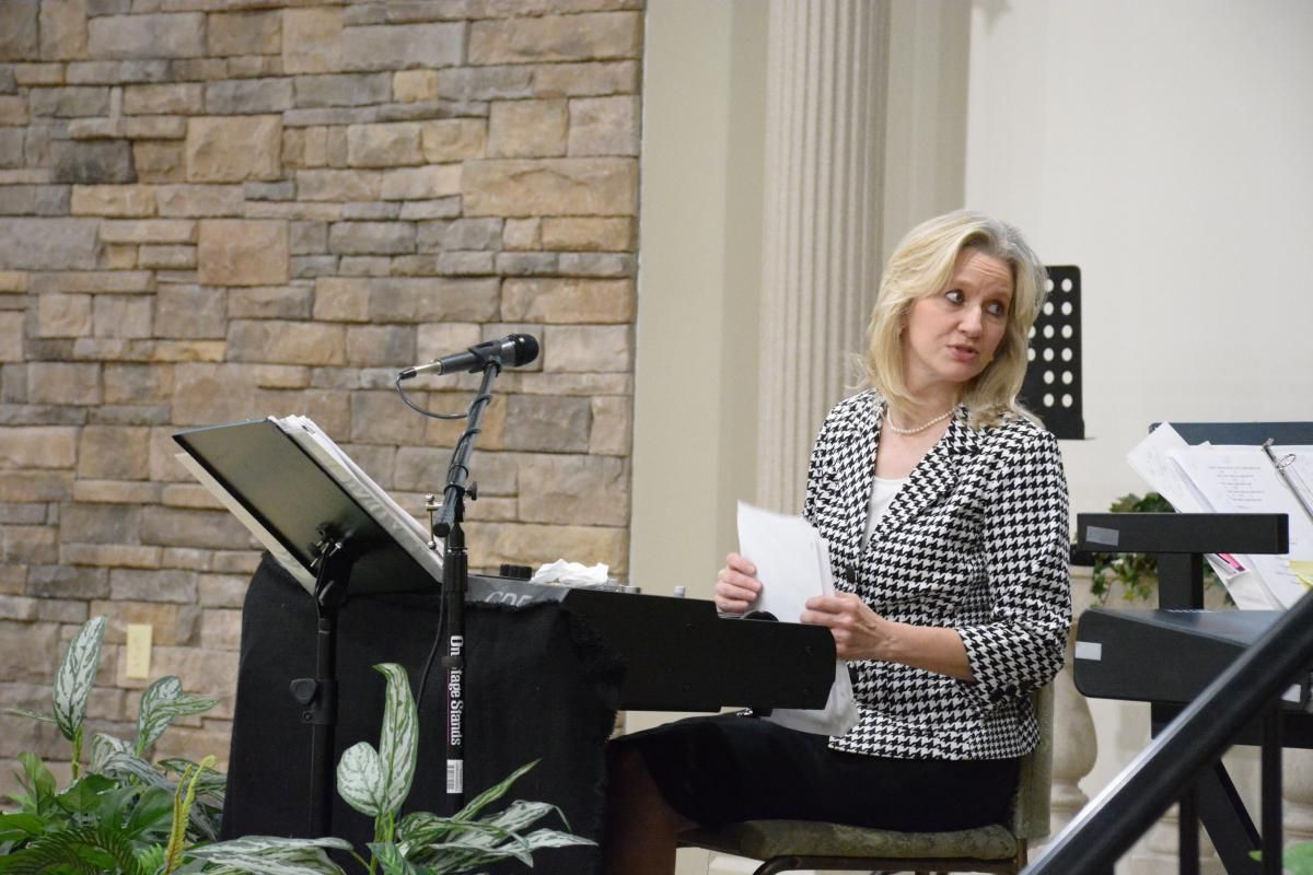 Woman in black and white jacket seated, holding papers, speaking at lectern with microphone.